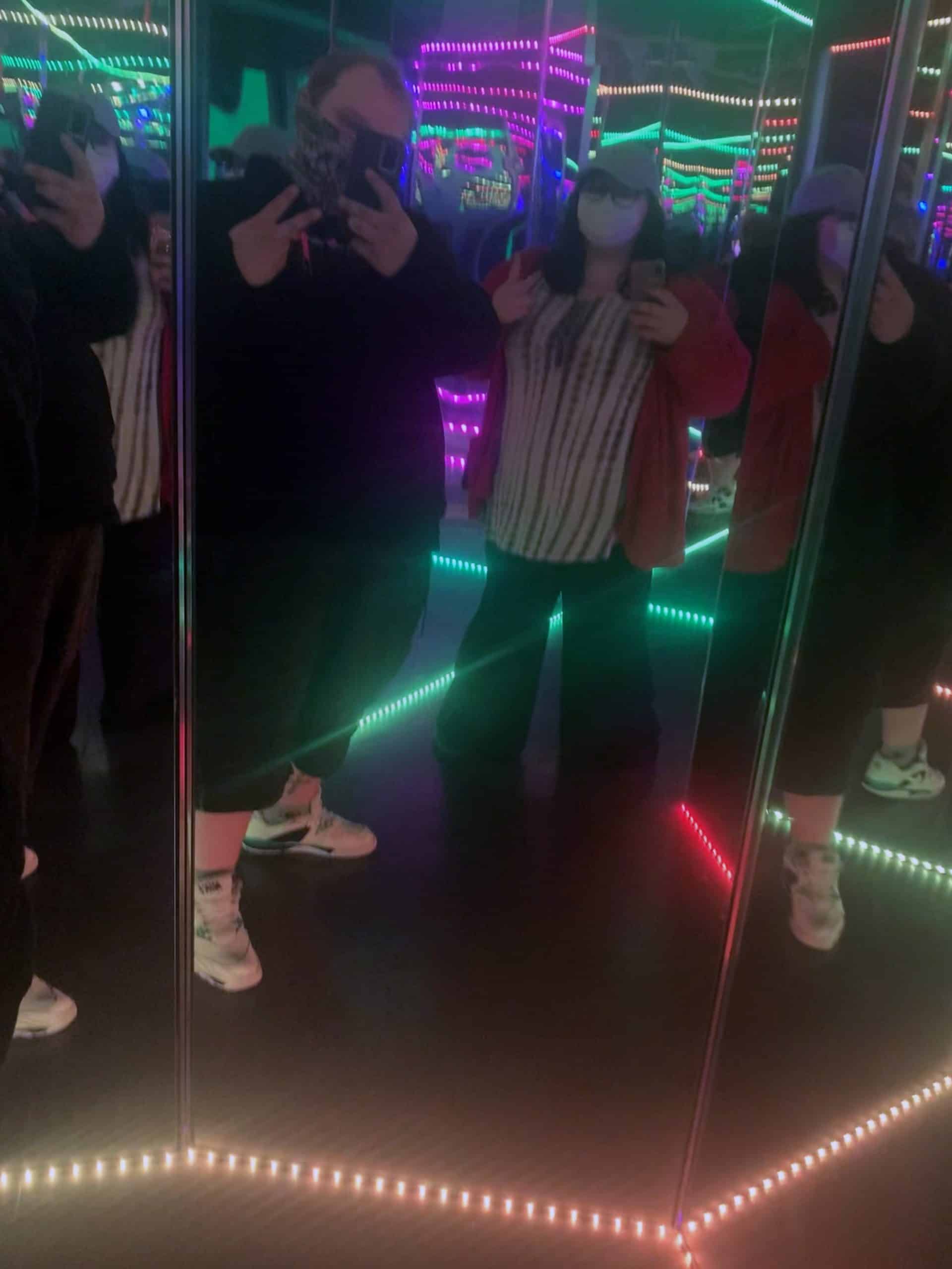 A man and woman standing in a mirrored hallway, brightlyy lined with rainbow LED lights