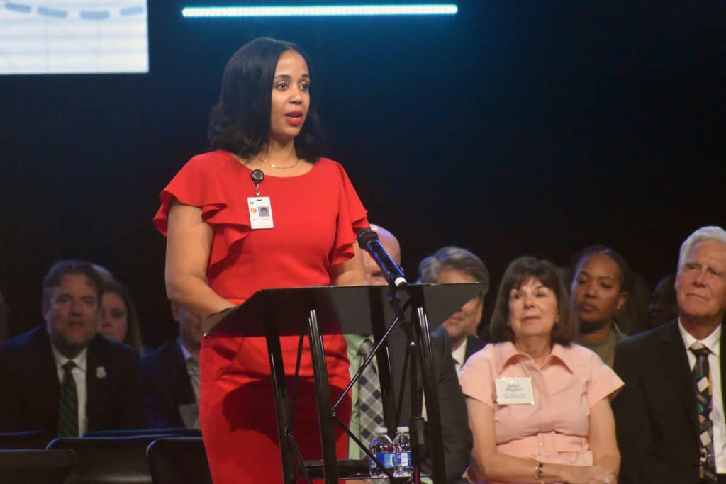 A woman in a red dress speaks at a podium, wearing a badge with her photo and name. Behind her, several people are seated and listening attentively. The background is dark, with some stage lights visible above.
