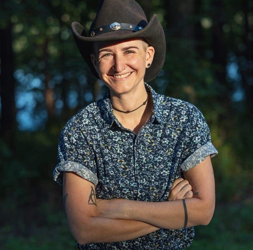Photo of a woman smiling for the camera. She is wearing a cowboy hat and crossing her arms.